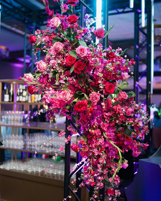 Large vibrant floral arrangement of pink and red roses with cascading smaller pink flowers, displayed indoors with bar shelves and glassware in the background.