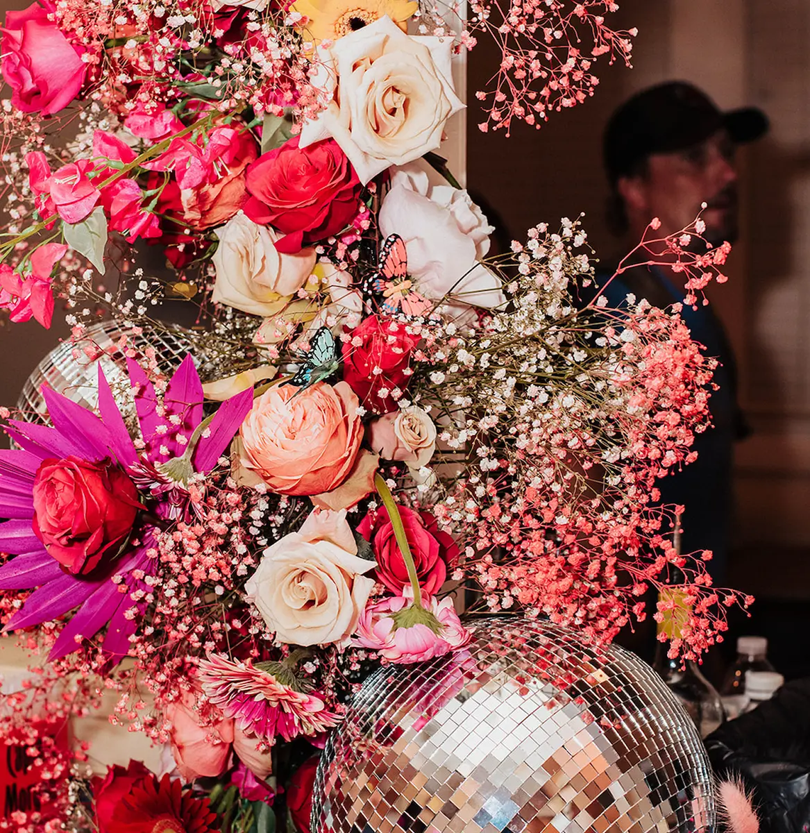 Colorful floral arrangement with roses, pink flowers, delicate baby's breath, two decorative butterflies, and a reflective disco ball with a blurred person wearing a cap in the background.