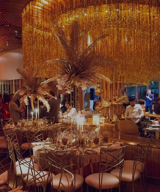 Elegant dining table set with tall feather centerpieces and lit candles in a warmly lit, gold-decorated venue with people in the background.