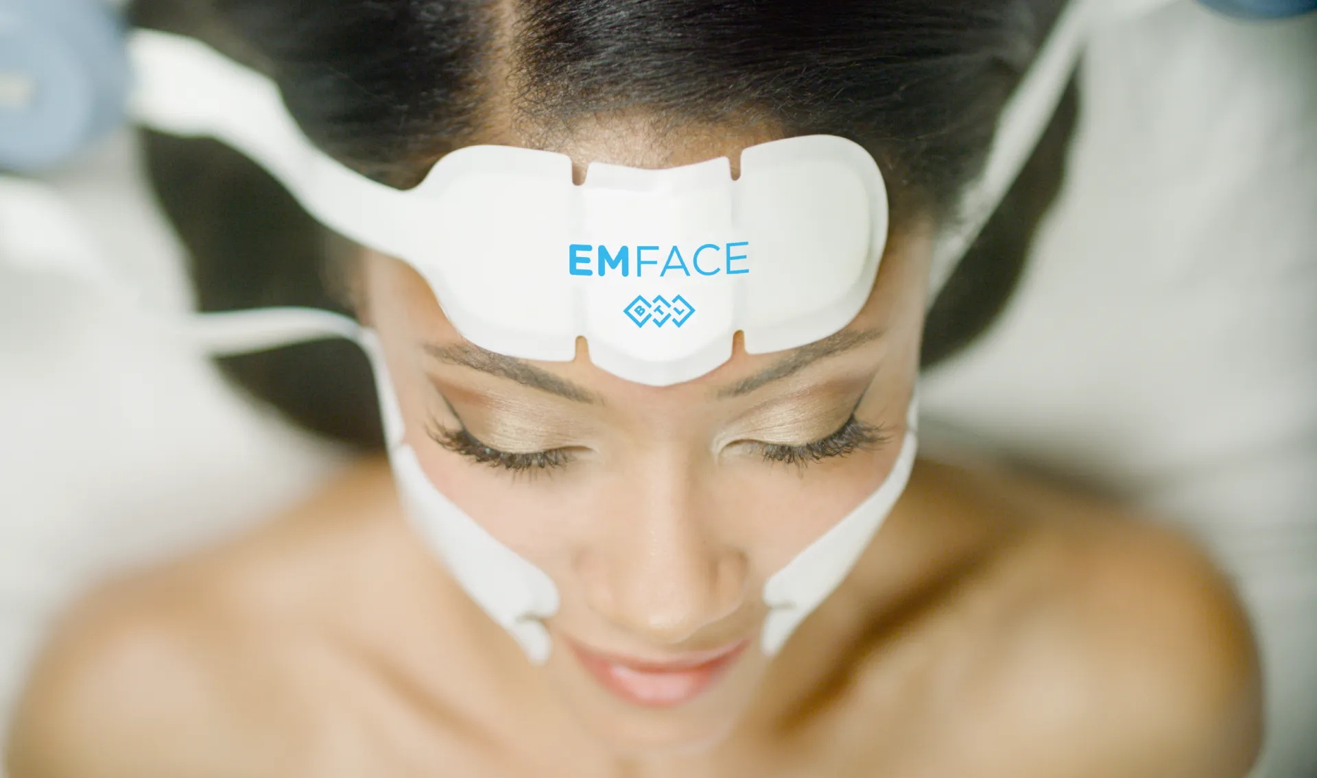 Top-down view of a woman lying down during a non-invasive EMFACE facial rejuvenation treatment, wearing specialized white applicator pads on her forehead and cheeks to lift muscles and reduce wrinkles.
