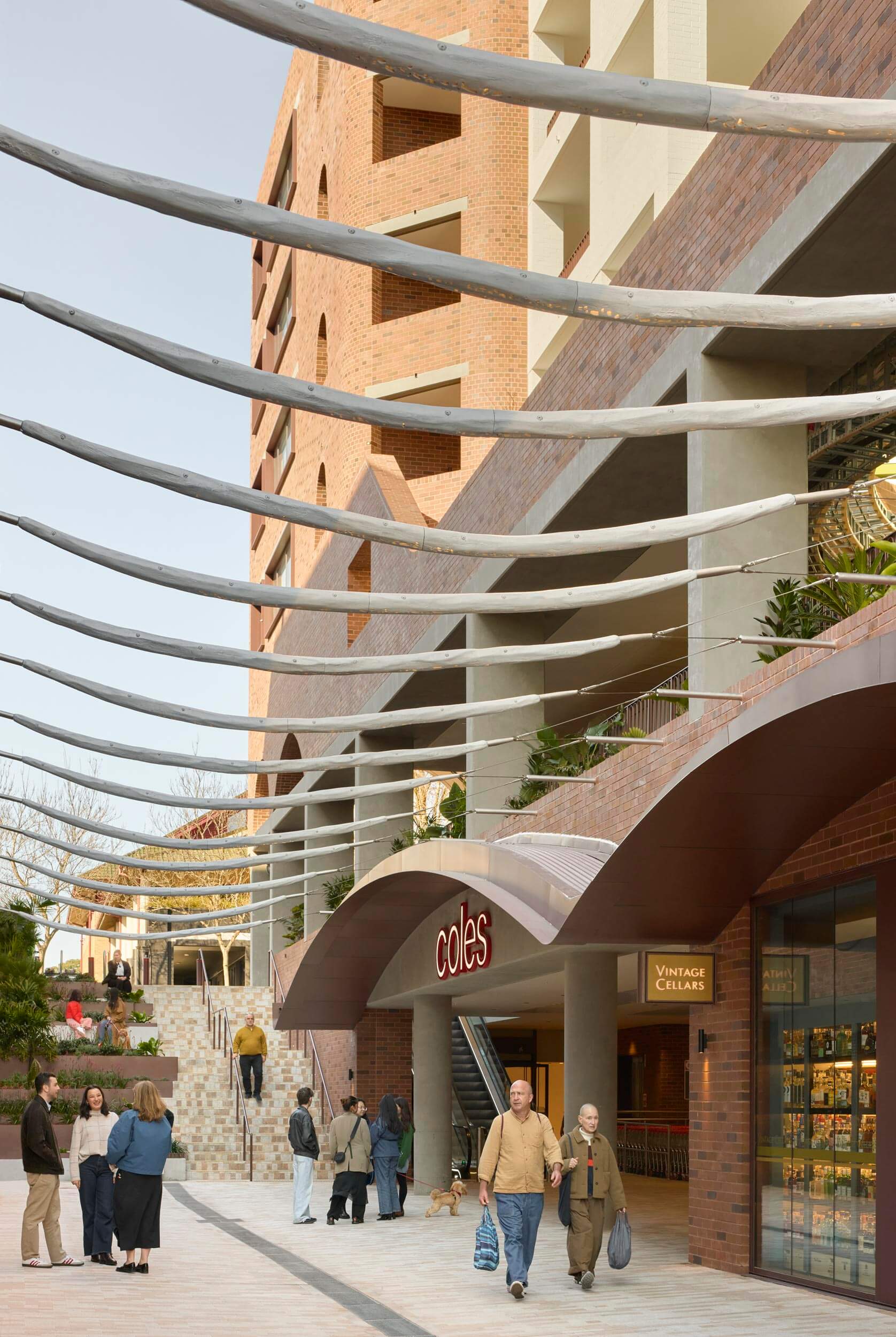 Couple walking past a modern residential building and supermarket in a landscaped public area.