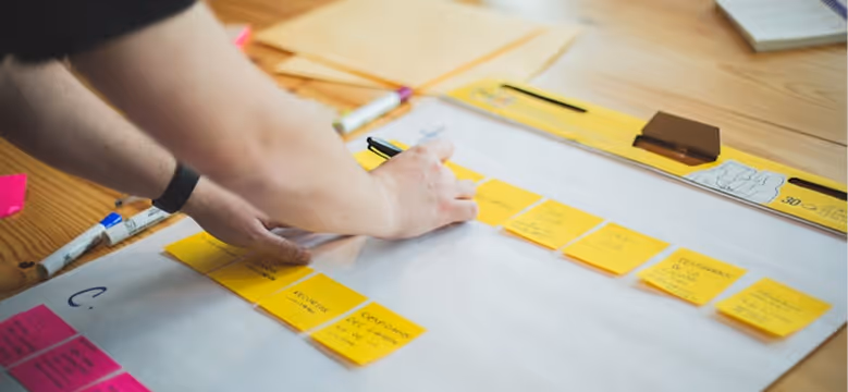 Person arranging and writing on yellow sticky notes placed in rows on a large white paper sheet on a wooden table.