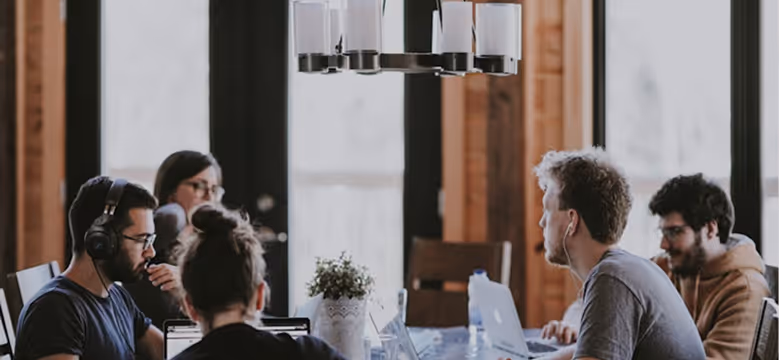 Group of five people working on laptops around a wooden table in a modern office with large windows.