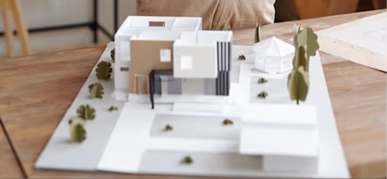 Architectural scale model of a modern house with trees on a white base placed on a wooden table.