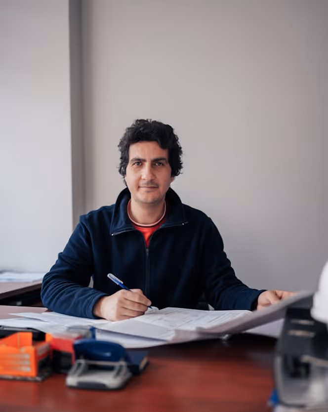 Man with curly hair wearing a dark jacket sitting at a desk writing on documents.
