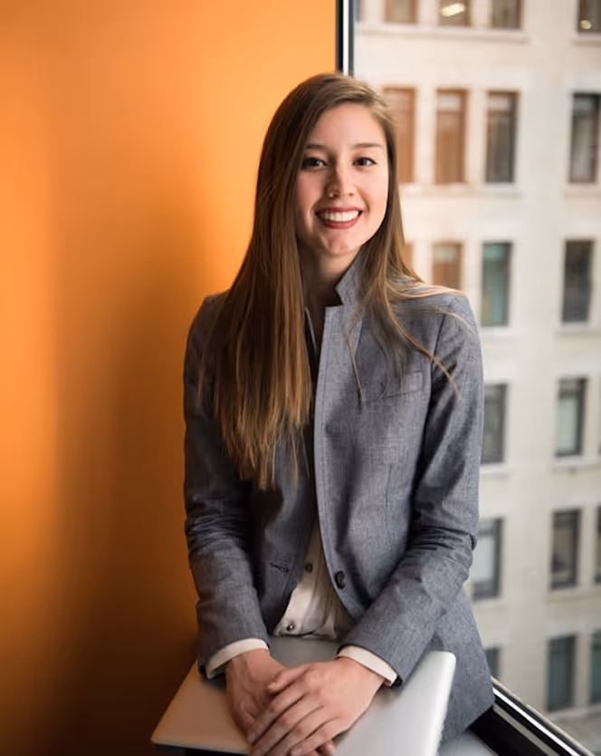 Smiling woman with long brown hair wearing a gray blazer sitting by a window holding a laptop.