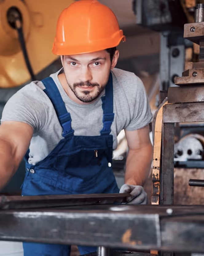 Male factory worker wearing an orange hard hat and blue overalls operating machinery and handling a metal piece.