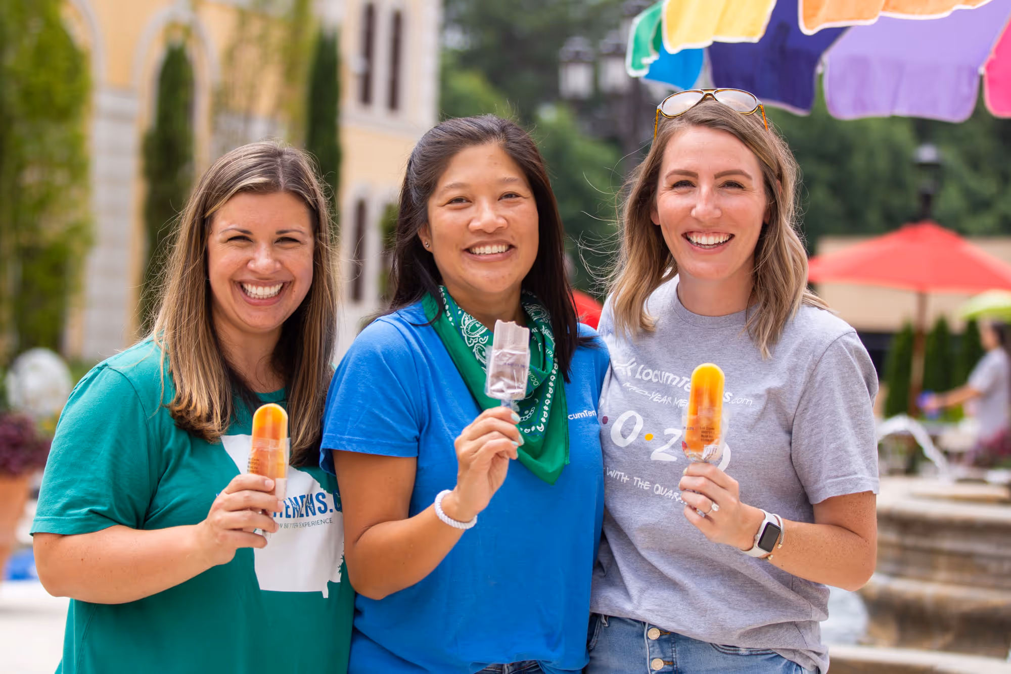 Three women smiling and holding popsicles at an outdoor community celebration with colorful umbrellas in the background.