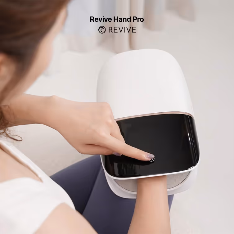 Mujer usando un masajeador de manos Revive Hand Pro, colocándose la mano en el dispositivo blanco con una pantalla negra.