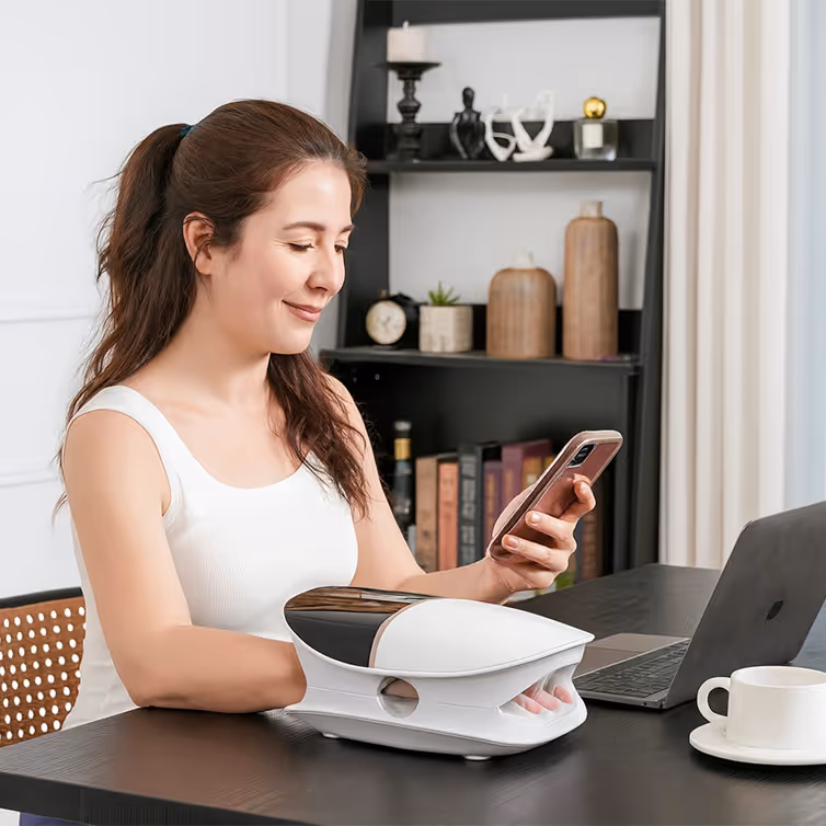 Mujer usando un dispositivo de terapia para la mano mientras mira su teléfono, sentada en un escritorio con una computadora portátil y una taza blanca.