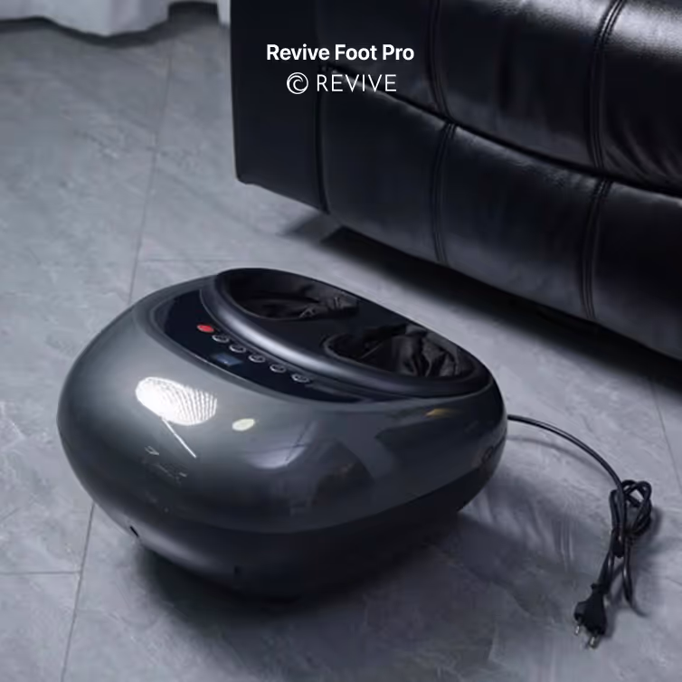 Black Revive Foot Pro foot massager placed on a gray tiled floor next to a black leather couch.