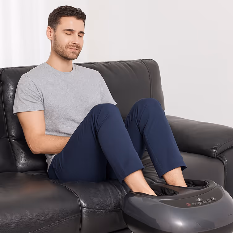 Hombre sentado en un sofá negro usando un masajeador eléctrico para pies con expresión de relajación.