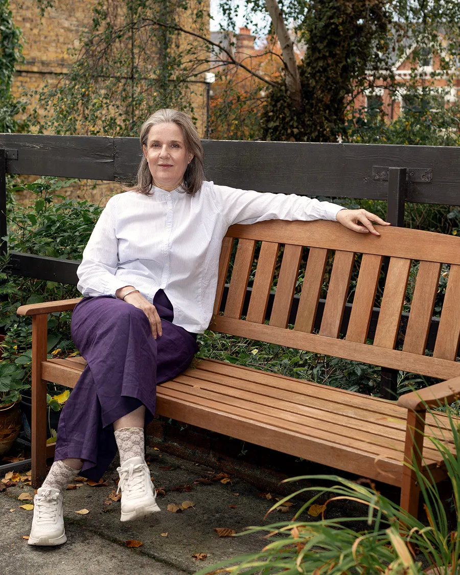 Sophrologist Lucile Dieblery sitting on a wooden bench with legs crossed wearing a white shirt and purple trousers