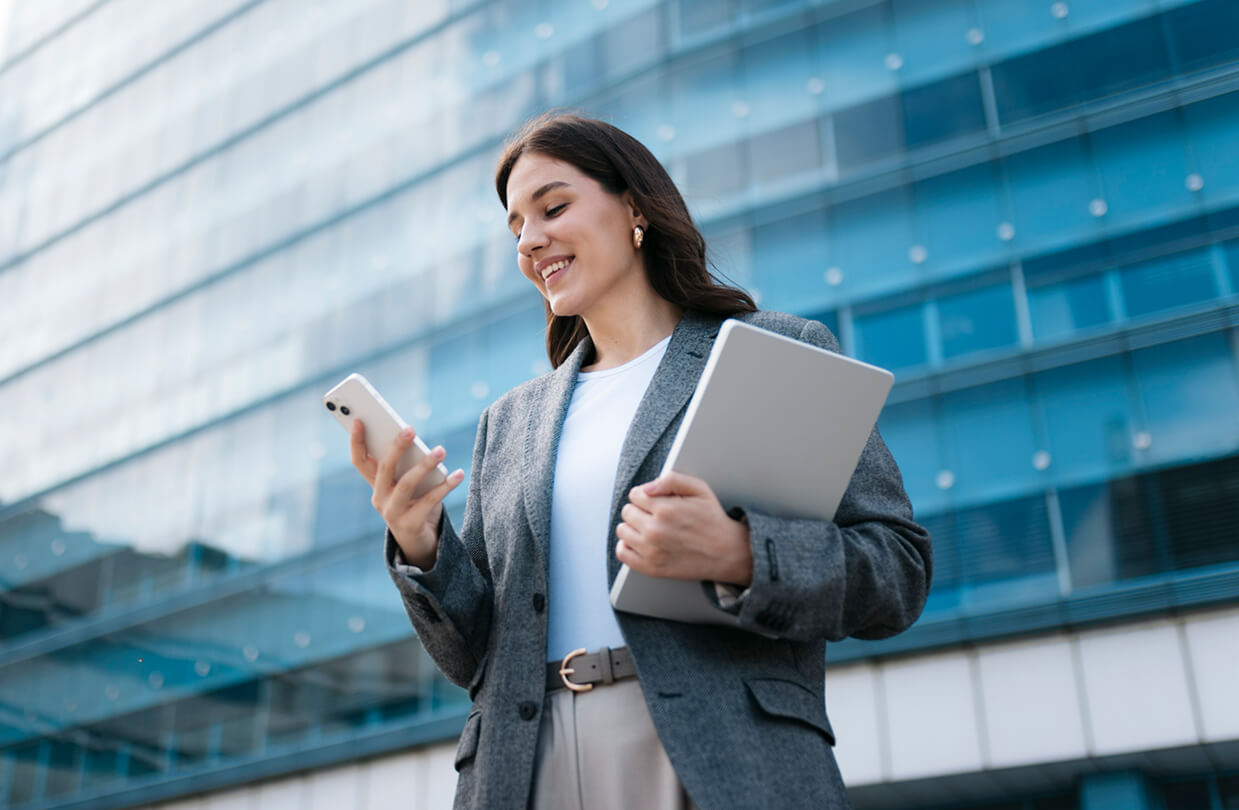 Walking looking at her phone holding a laptop with a building behind her