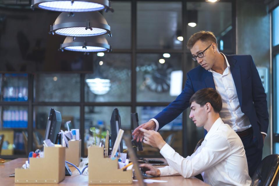 Two men look at information on a desktop computer screen. One man sits at the desk, the other stands over his shoulder, gesturing to the screen.