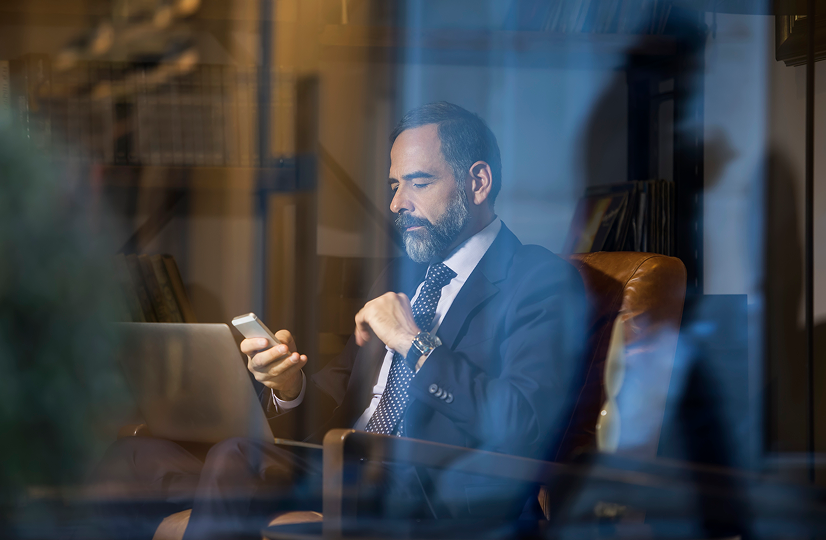 Photo taken through a window with reflections on it. Inside the window, a man sits in an armchair and reads on his phone.