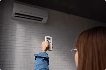 Person holding a remote control pointed at a wall-mounted air conditioner unit.