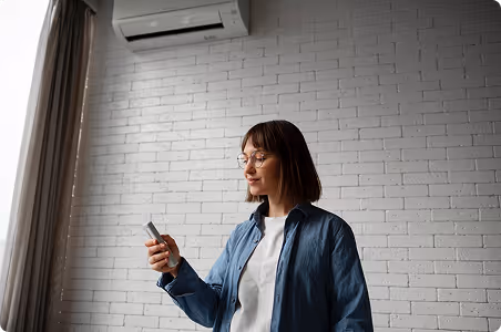 Woman holding a remote control, standing indoors in front of a white brick wall and an air conditioner.