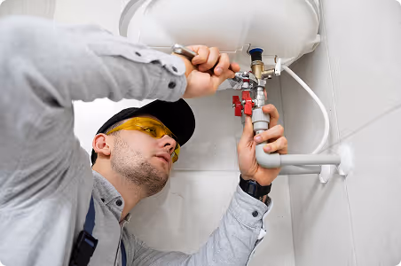 Man wearing safety glasses and cap fixing a pipe under a water heater with a wrench.