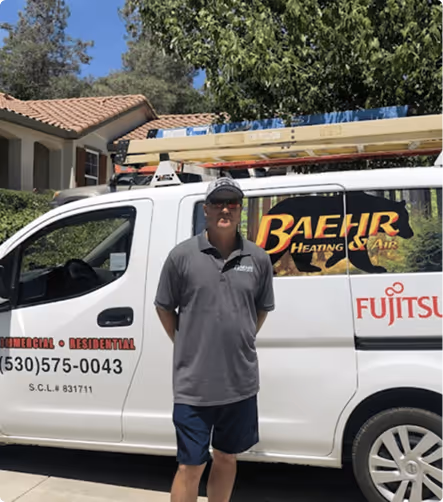 Man wearing sunglasses, a gray polo shirt, and navy shorts standing in front of a white work van with BAEHR Heating & Air logo and Fujitsu branding, parked outside a house with trees in the background.