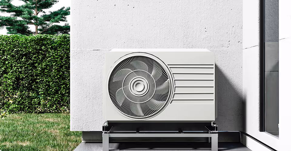White outdoor air conditioning unit mounted on metal stand outside a white building wall with greenery in the background.