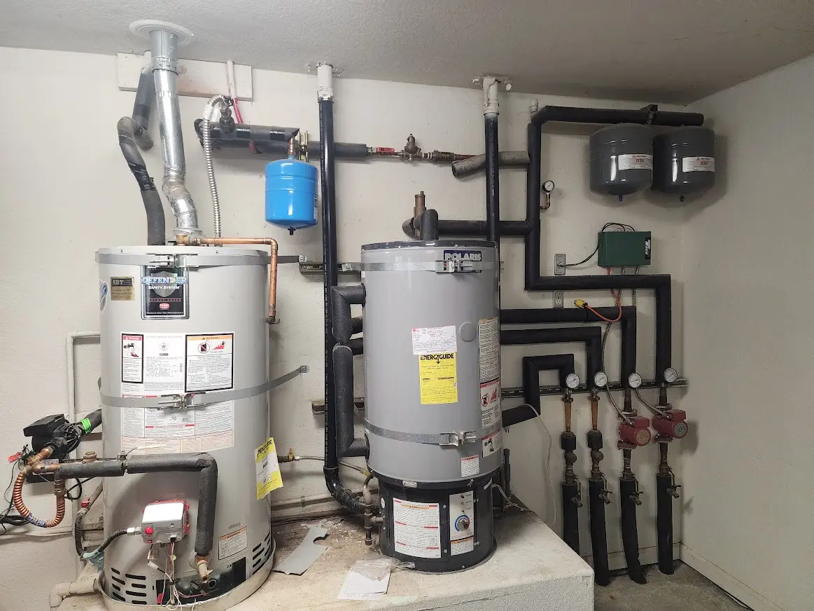 Two industrial water heaters connected to a network of insulated black pipes, pressure gauges, and valves mounted on a wall.