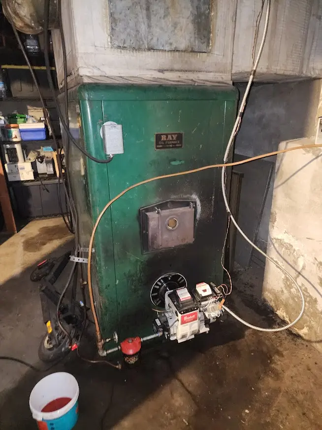 Old green RAY oil furnace in a basement with attached burner and visible pipes and wires.