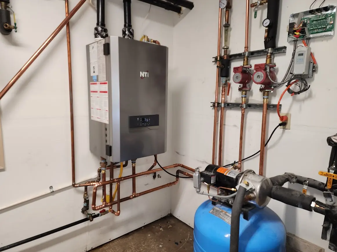 Utility room with a wall-mounted NTI tankless water heater, copper piping, pressure gauges, two red pump valves, and a blue pressure tank.