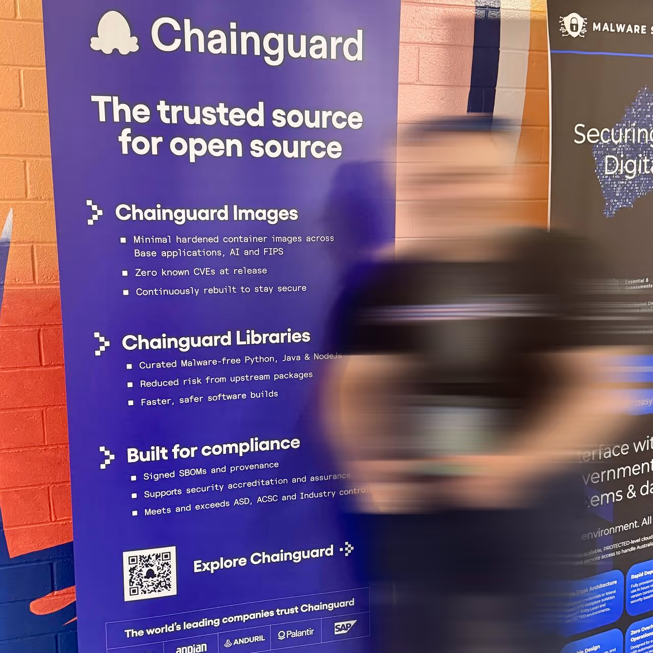 Smiling man in a black Chainguard t-shirt giving thumbs up next to a purple informational banner about Chainguard's open source security solutions.