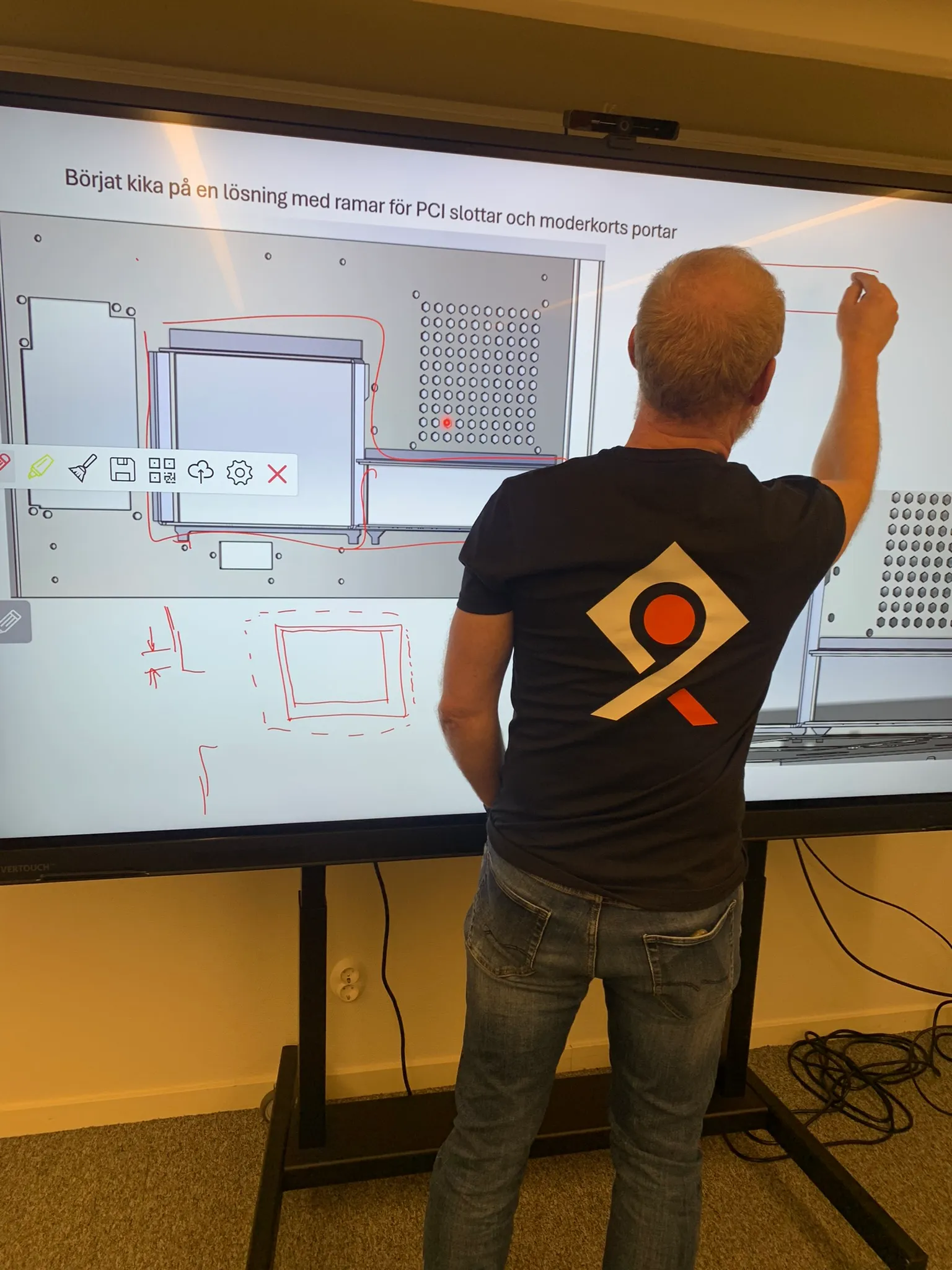 Man in black t-shirt with a geometric logo standing and drawing on a large touchscreen displaying a technical diagram with red annotations.