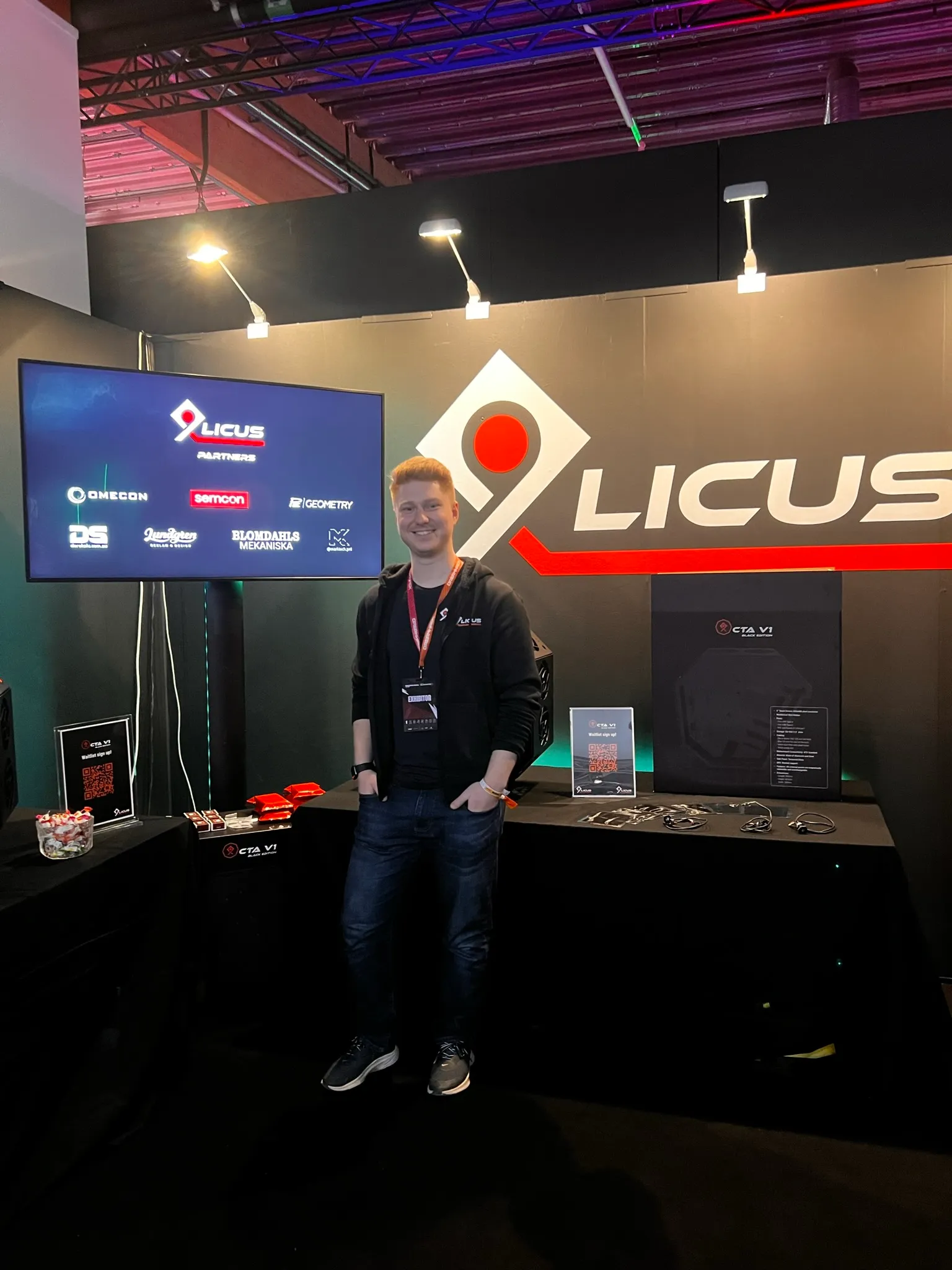 Man standing in front of a Licus exhibition booth with a large logo on the wall and a screen displaying partner logos.
