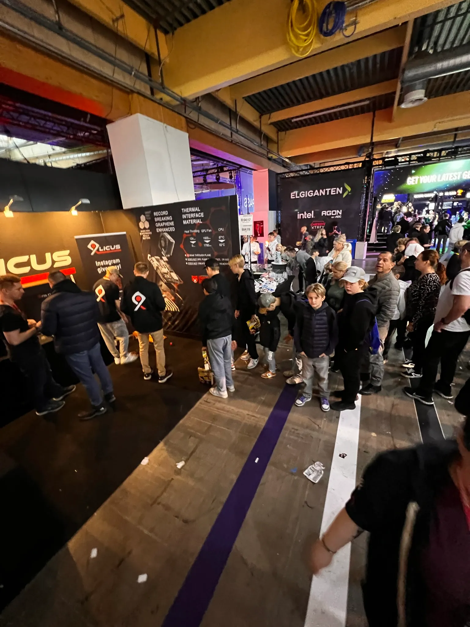 Crowd of people and children standing in line inside a convention or gaming event with several branded booths including 9licus and Elgiganten.