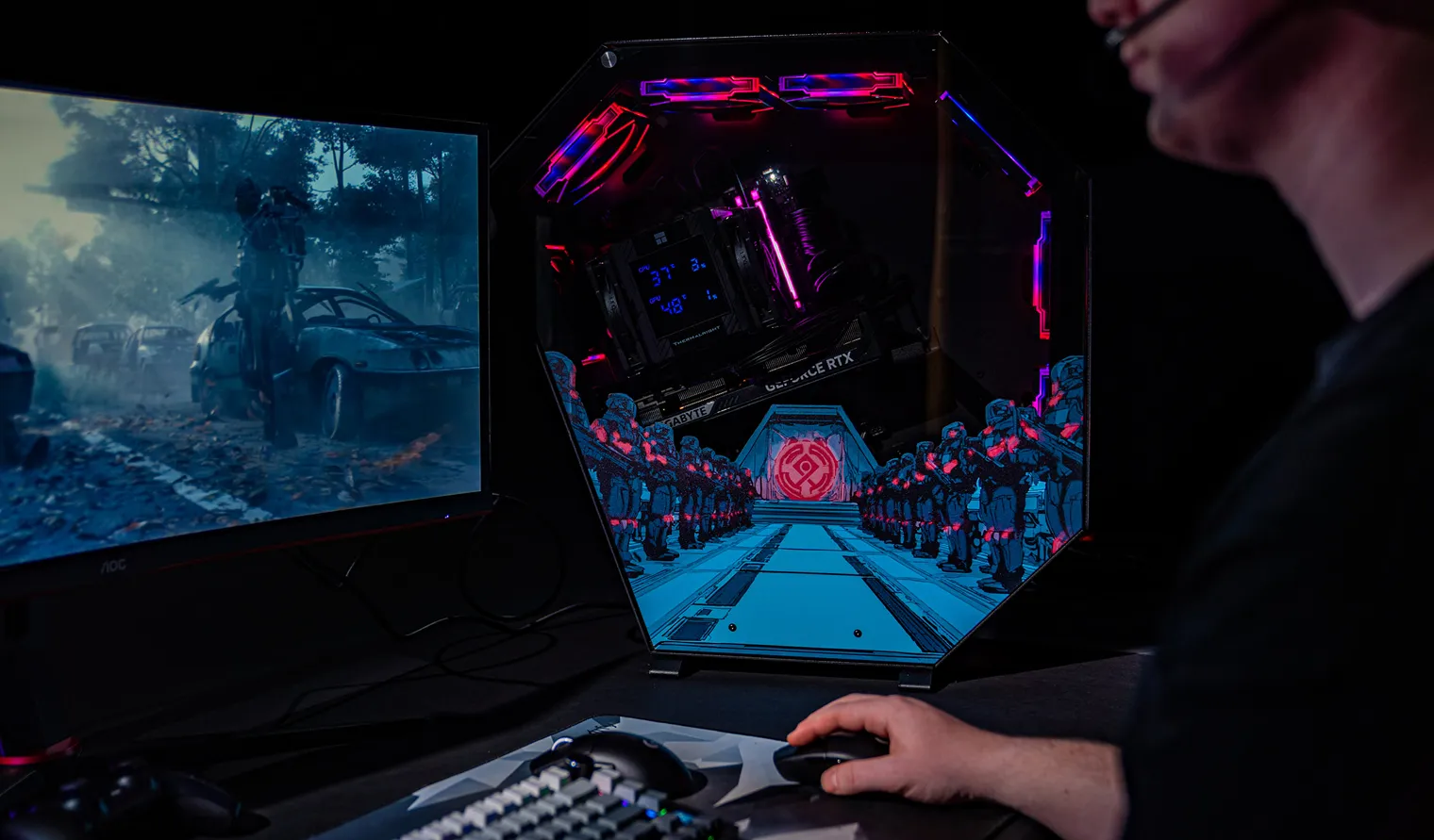 Person gaming on a PC setup showing a post-apocalyptic scene on the monitor and colorful LED-lit PC components inside a polygonal case.