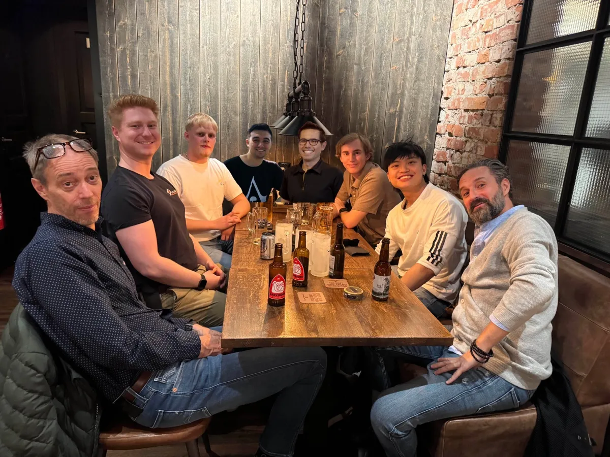 Eight men sitting around a wooden table in a cozy restaurant with drinks and glasses in front of them.