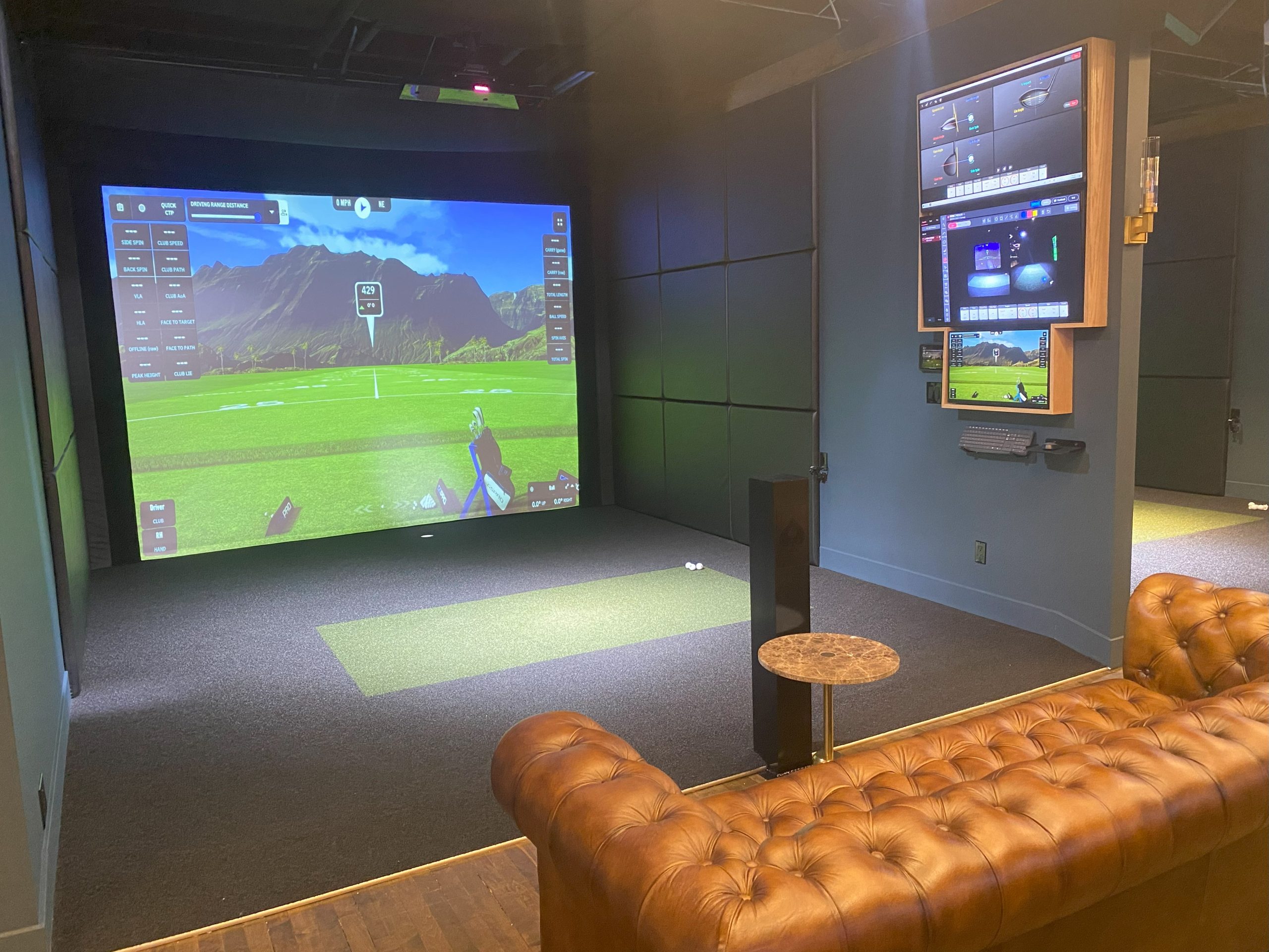 Indoor golf simulator room with a large screen displaying a golf game, a putting green area, brown leather tufted sofa, small round table, and monitors mounted on the wall.