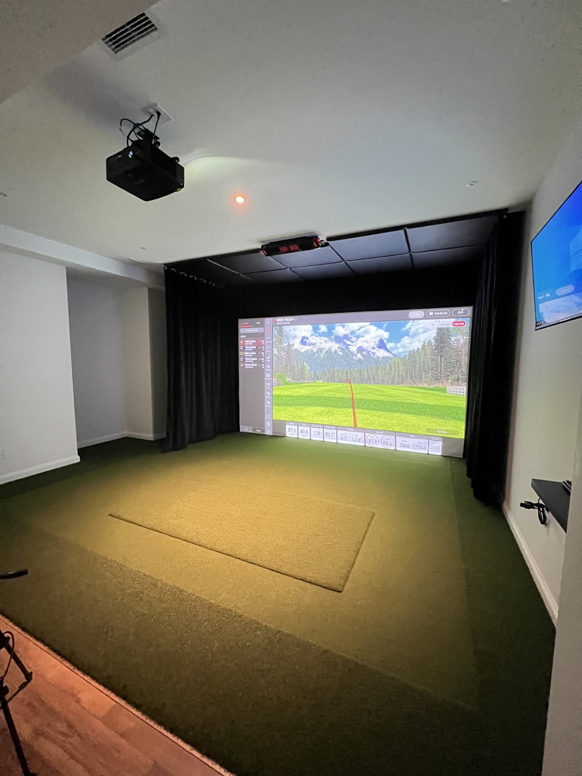 Indoor golf simulator room with projector screen showing a virtual golf course and artificial turf flooring.