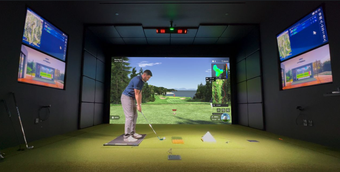 Man preparing to hit a golf ball in an indoor golf simulator room with large screen showing a golf course and two side monitors displaying game data.