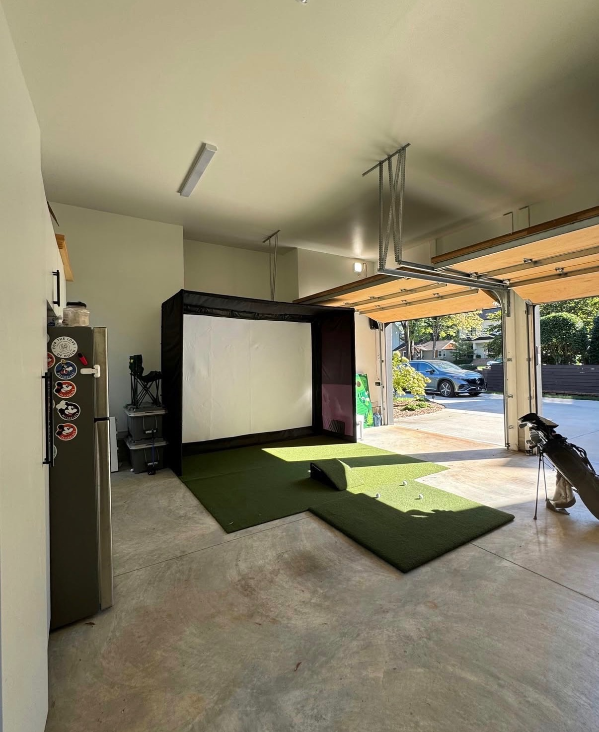 Indoor golf simulator setup in a garage with artificial turf, golf balls, and clubs, and an open garage door showing a car outside.