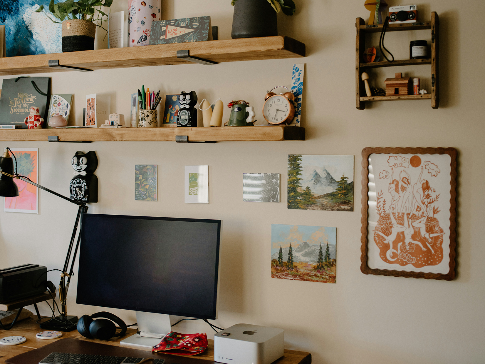 Home office desk with a computer monitor, keyboard, headphones, and artwork on the wall, including paintings and framed prints.