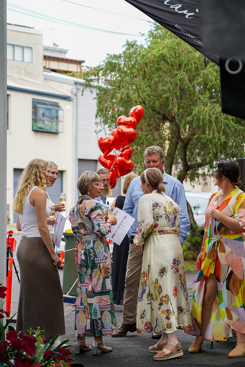 Group of six people standing and chatting outdoors near red heart-shaped balloons, some holding wine glasses and papers, dressed in colorful and patterned clothing.