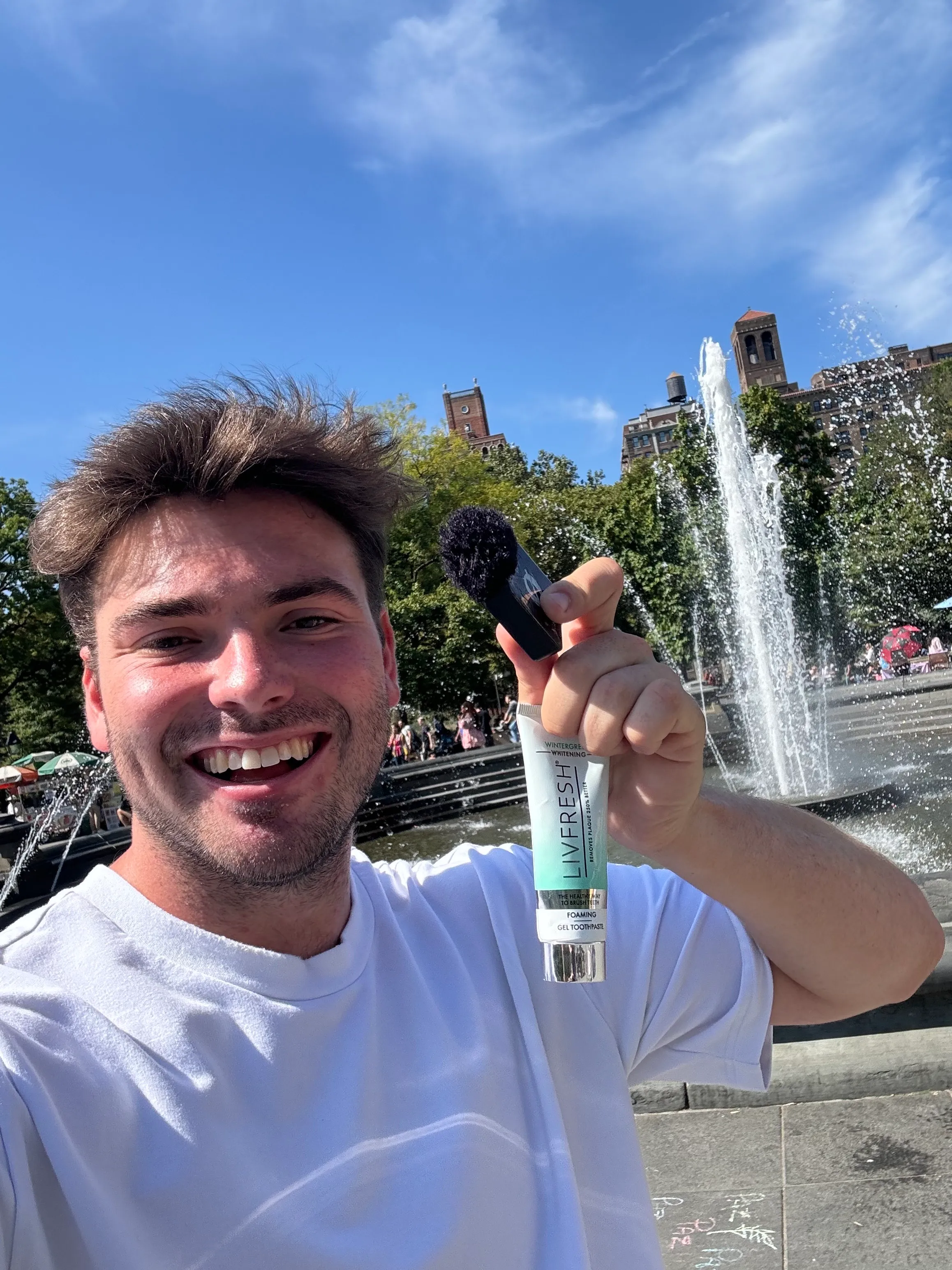 Smiling man in a white shirt holding a tube of Liverfresh whitening toothpaste and a toothbrush near a fountain in a sunny park.