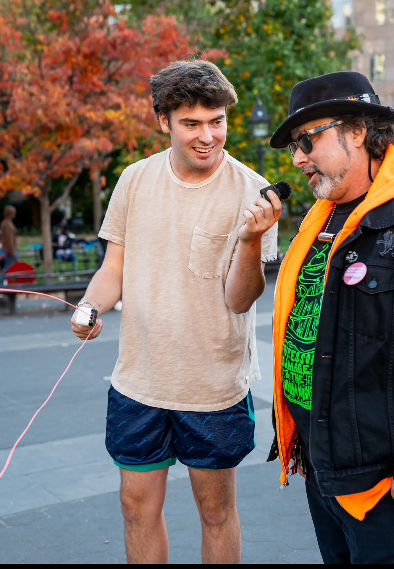 A young man in shorts and a t-shirt holds a microphone towards a man wearing a black hat, sunglasses, and a neon orange jacket in a park setting with autumn trees.