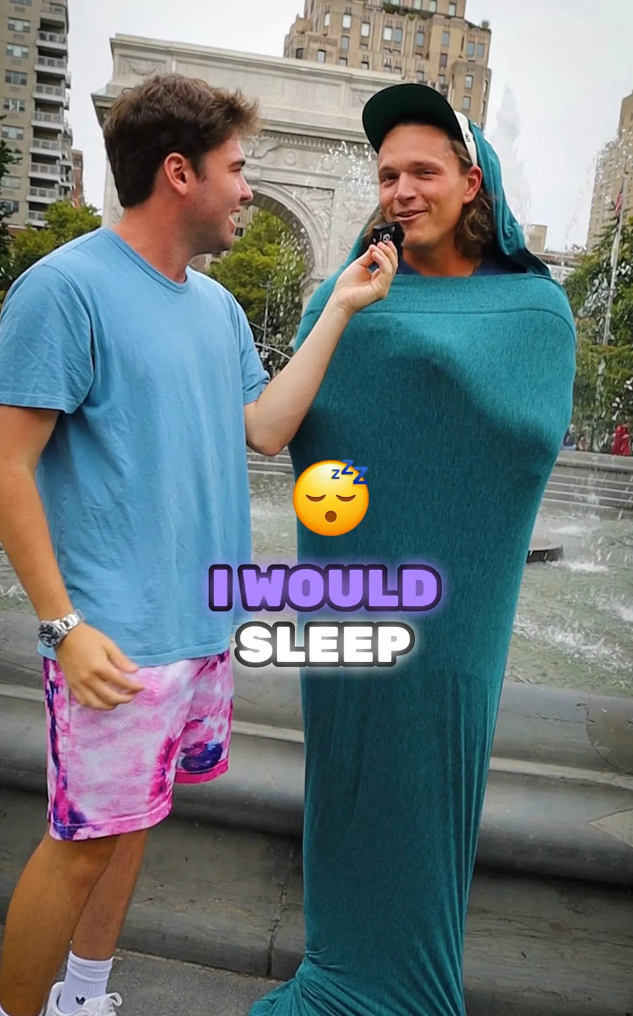 Man in blue shirt interviews another man wrapped in a teal fabric standing near a fountain with buildings in the background.