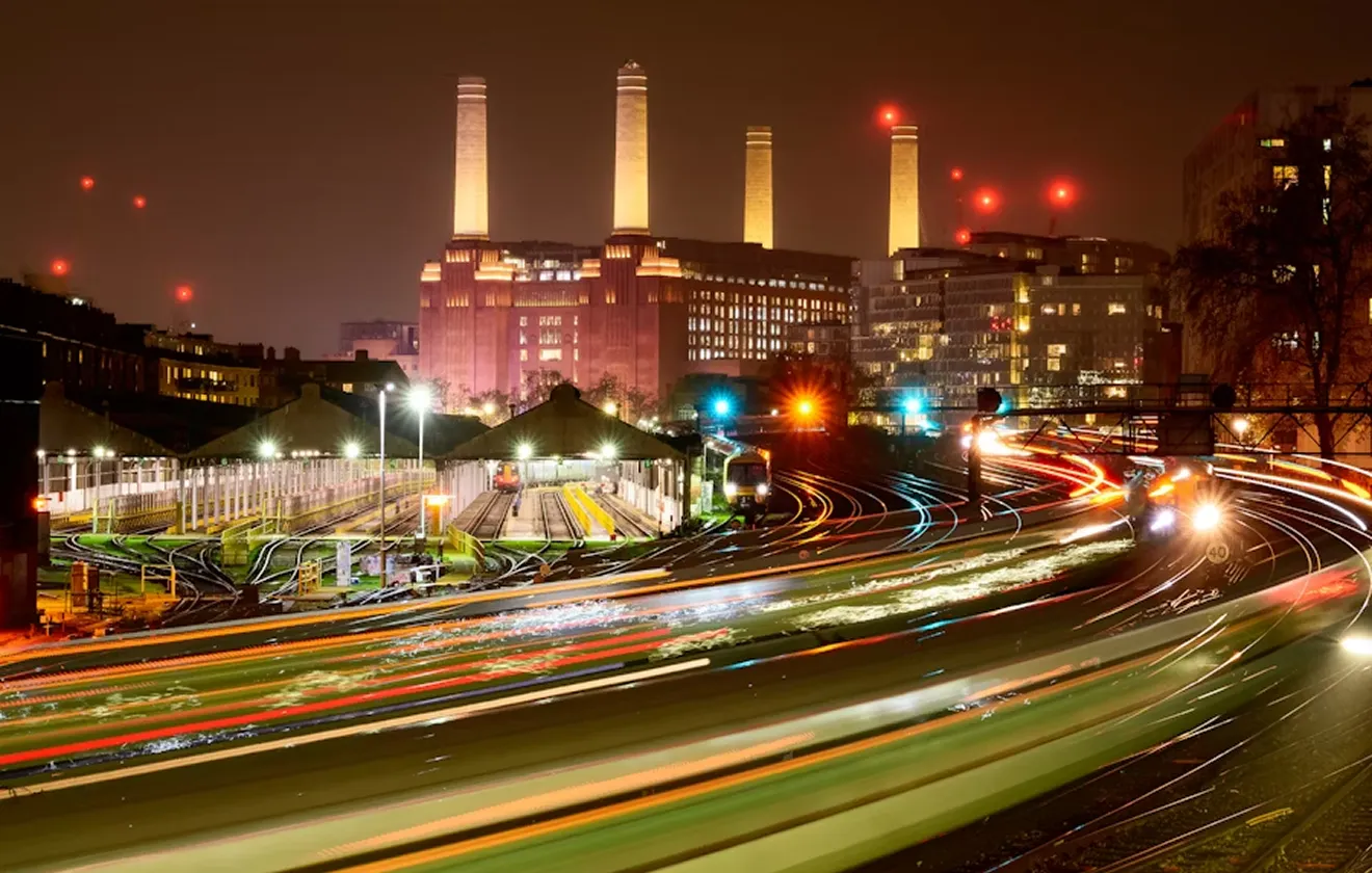 Night view of an illuminated industrial building with four tall chimneys behind a busy railway yard with light trails from moving trains.