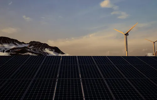 Solar panels in the foreground with wind turbines and mountains under a partly cloudy sky at sunset.