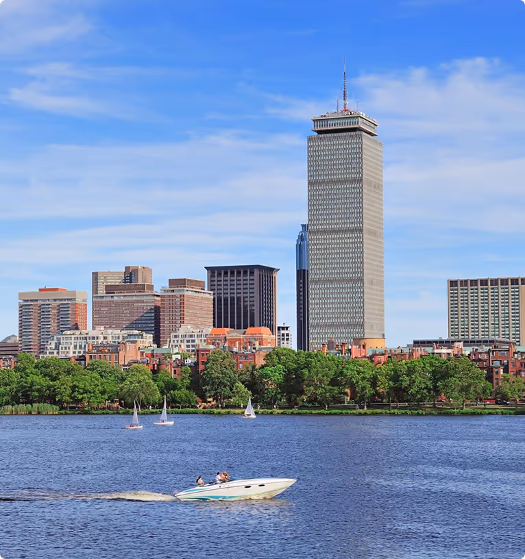 City skyline with tall buildings behind a tree-lined riverbank and a speedboat and sailboats on the water under a partly cloudy blue sky.