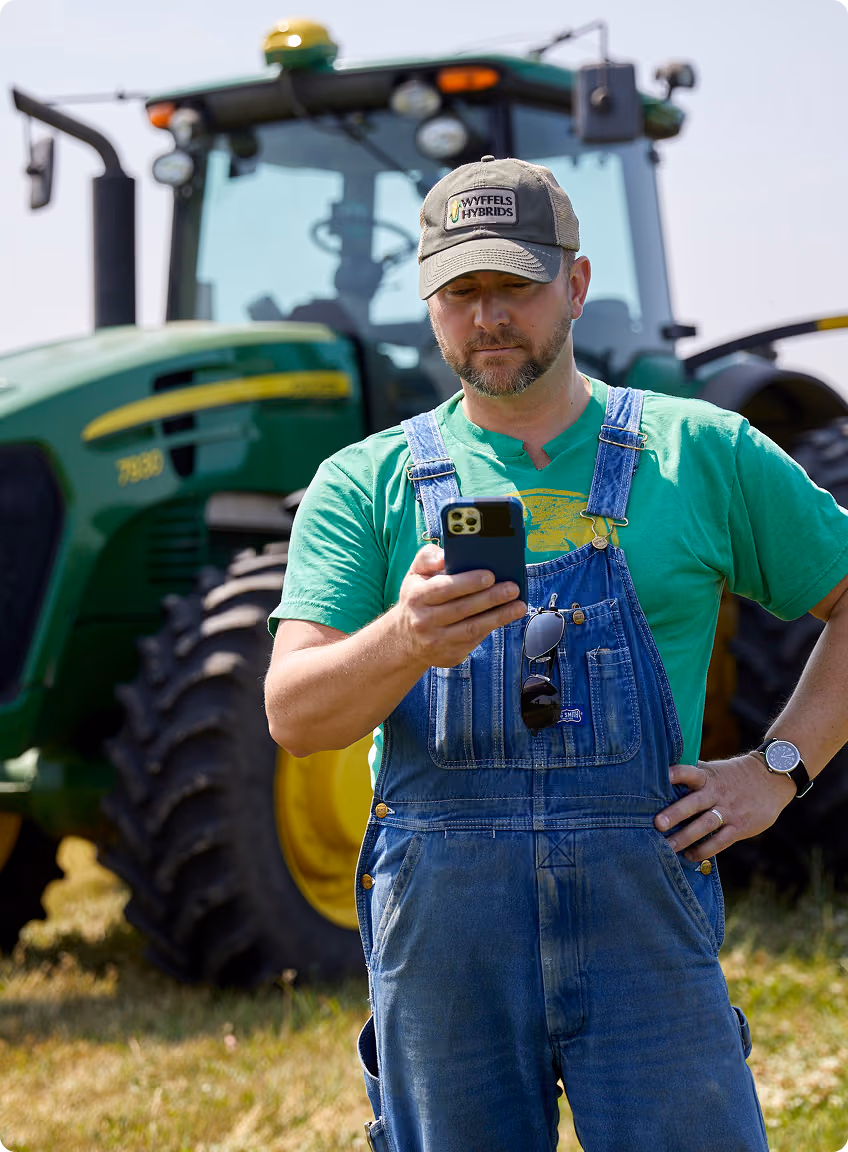 Farmer in green shirt and denim overalls looking at smartphone in front of a green tractor.