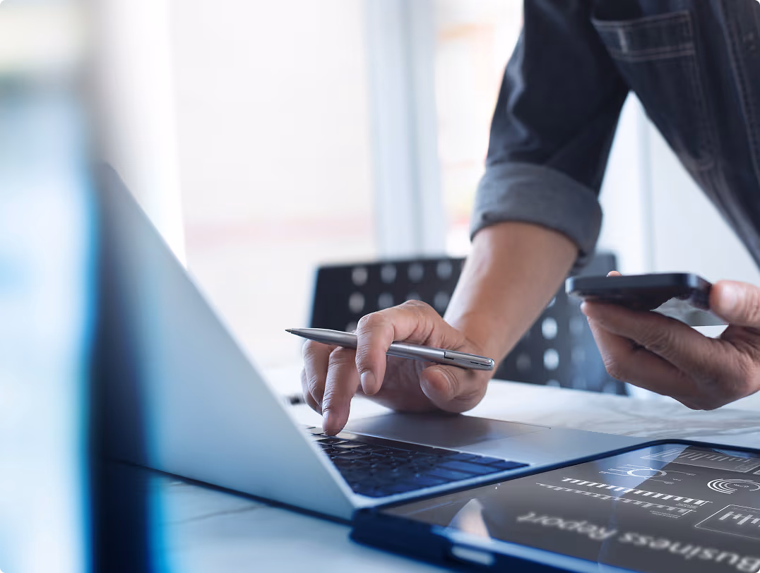 Person using a pen to interact with a laptop keyboard while holding a smartphone, with a tablet displaying business report charts on a desk.