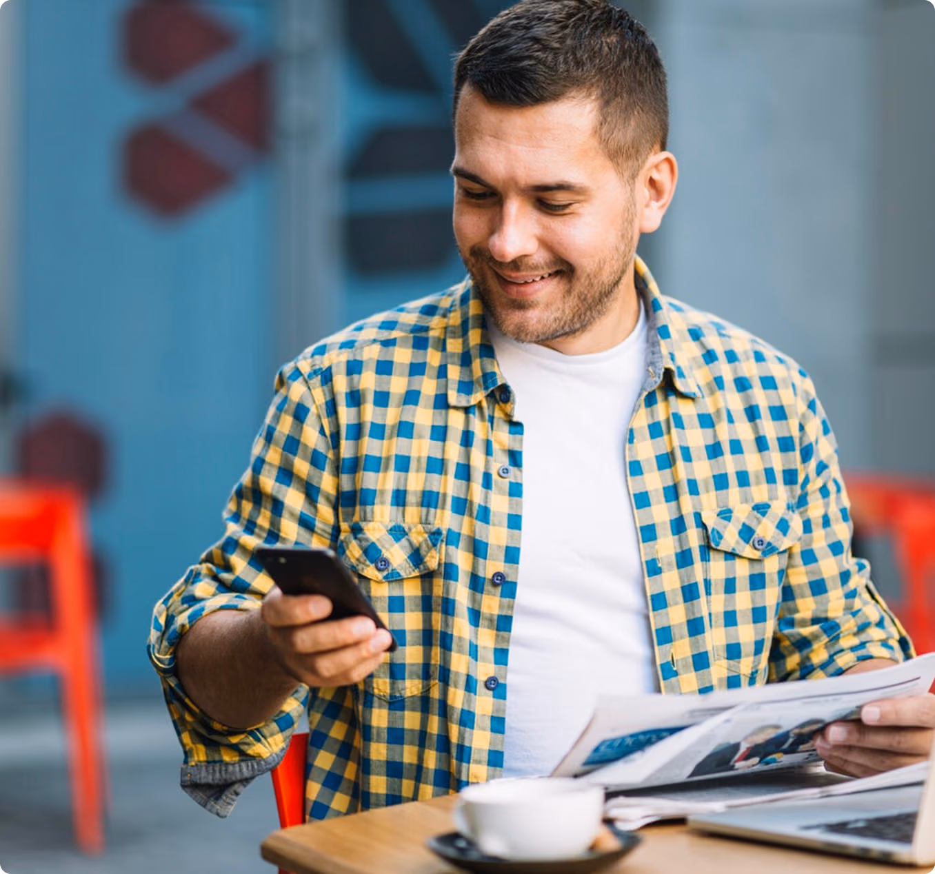 Smiling man in yellow and blue plaid shirt holding a smartphone and reading a newspaper at a table with a cup of coffee and laptop.