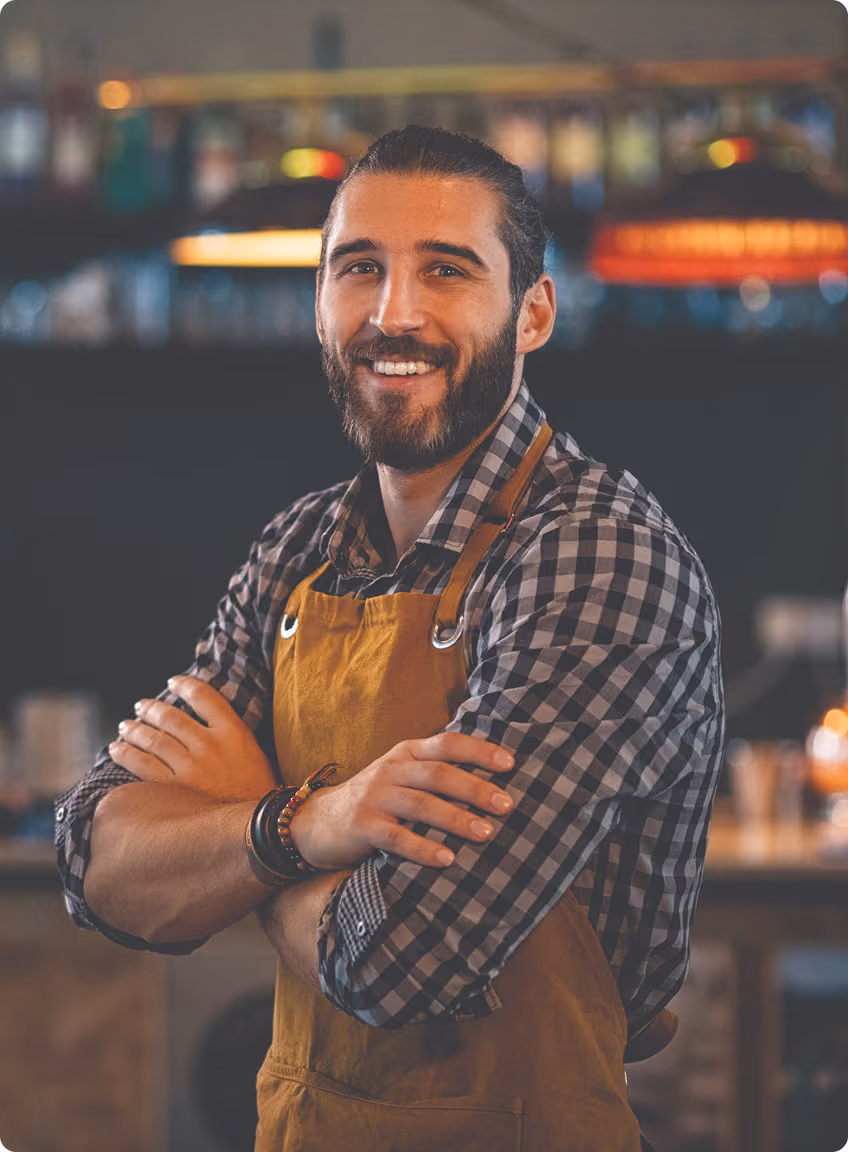 Smiling bearded man with a bun wearing a checkered shirt and mustard apron with arms crossed in a cozy café interior.
